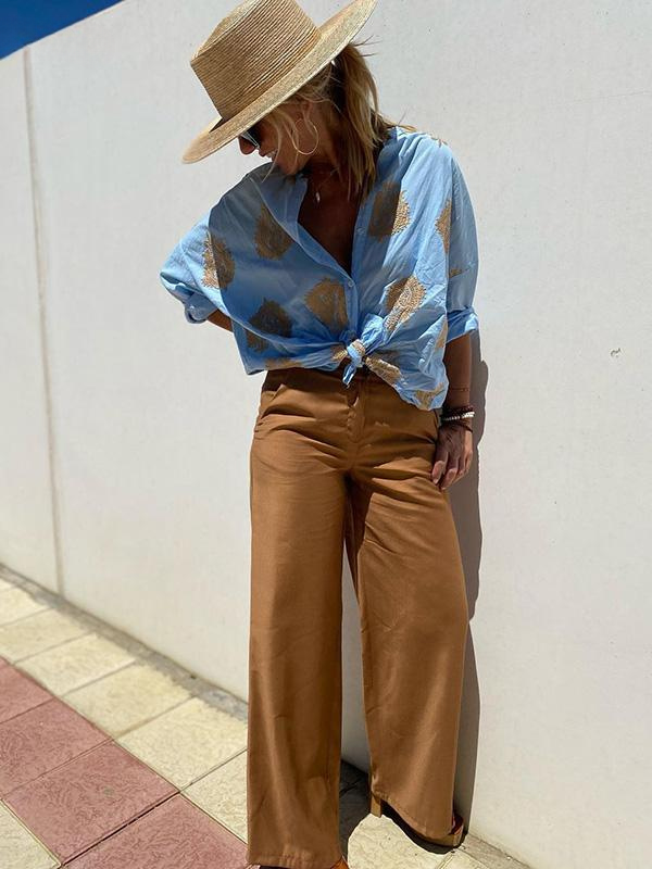 Frau in blauer Bluse mit goldenen Mustern, braune Hose, Strohhut, Sommermode, lässiger Stil.