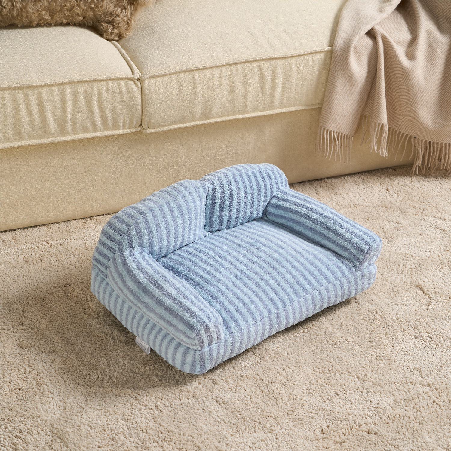 Gestreiftes blaues Haustiersofa aus Stoff auf beigem Teppich vor einem cremefarbenen Sofa.