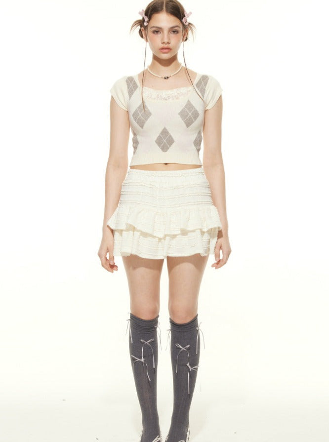 Junge Frau in modischem, weißem Outfit mit Rautenmuster-Top und Rüschenrock, trägt knielange Socken. Trendige Damenmode, stilvoll und modern.