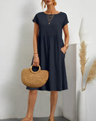 Frau in dunkelblauem Sommerkleid mit Strohtasche, goldenen Accessoires, stehend vor weißer Wand.