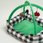 Schwarz-weiß kariertes Katzenbett mit grünem Plüschboden und hängendem Spielzeug.