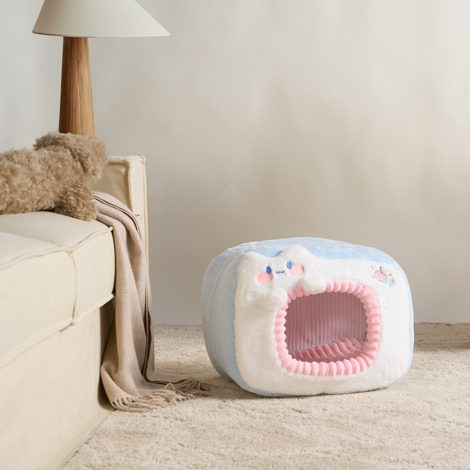 Katzenbett aus Plüsch in Blau und Weiß, quadratisch, mit rosa Öffnung, neben Sofa auf Teppich.