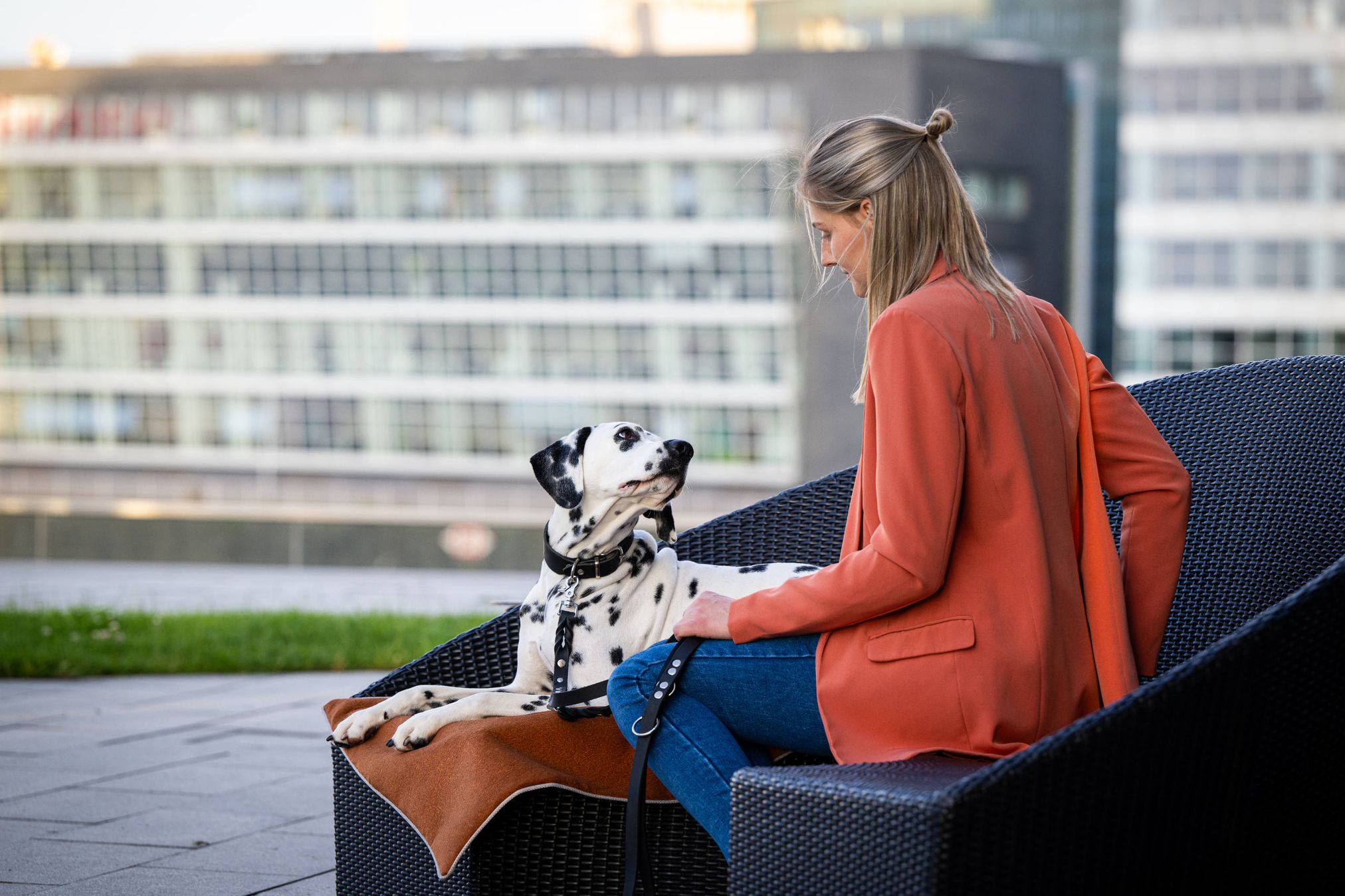Frau in rotem Blazer sitzt mit Dalmatiner auf schwarzem Outdoor-Sofa, moderne Stadtumgebung.