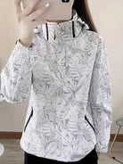 Weiße Damenjacke mit tropischem Blattmuster, Kapuze und Reißverschlusstaschen. Modische Outdoor-Bekleidung, ideal für Frühling und Herbst.