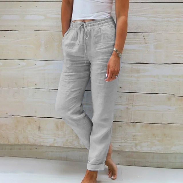 Frau in grauer Leinenhose und weißem Top vor Holzwand, lässige Sommermode.