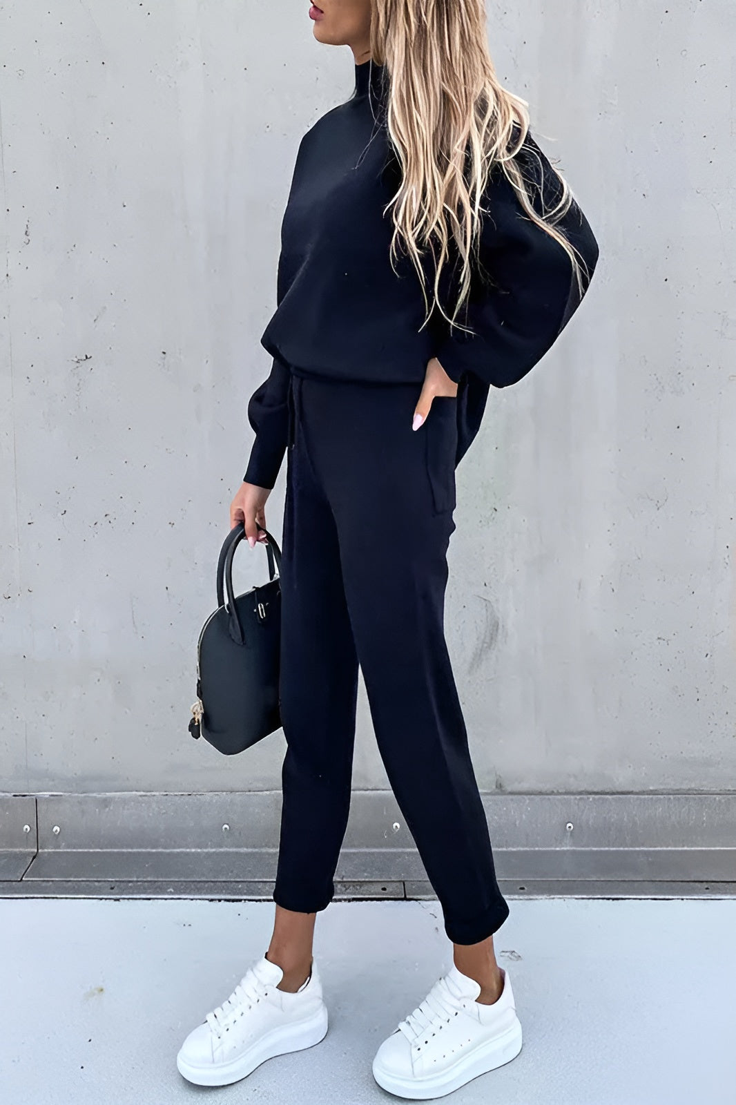 Frau in schwarzem Jogginganzug mit weißen Sneakers und schwarzer Handtasche vor grauer Wand. Modetrend, Freizeitlook, Damenbekleidung.