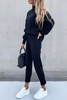 Frau in schwarzem Jogginganzug mit weißen Sneakers und schwarzer Handtasche vor grauer Wand. Modetrend, Freizeitlook, Damenbekleidung.