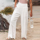 Frau in weißen Leinenhosen am Strand, hält eine Wasserflasche. Sommermode, Strandkleidung, lässiger Stil, bequeme Damenhosen.