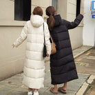 Zwei Frauen in langen Daunenmänteln, einer weiß und einer schwarz, spazieren auf einem Gehweg. Wintermode, warme Mäntel, stilvolle Outdoor-Bekleidung.