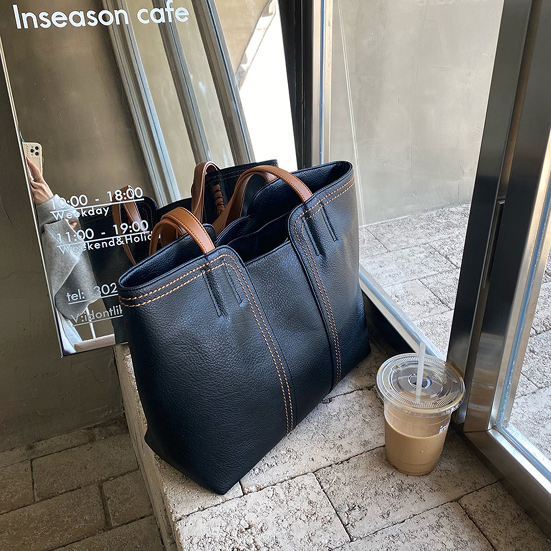Schwarze Ledertasche mit braunen Griffen auf Fensterbank, neben Eiskaffee. Stilvolle Handtasche, ideal für Alltag und Büro. Hochwertiges Accessoire.