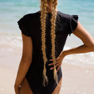 Frau in schwarzem Badeanzug am Strand, lange blonde Zöpfe, Sommermode, Strandbekleidung, stilvoller Einteiler, Urlaub, Meer, Bademode-Trends.