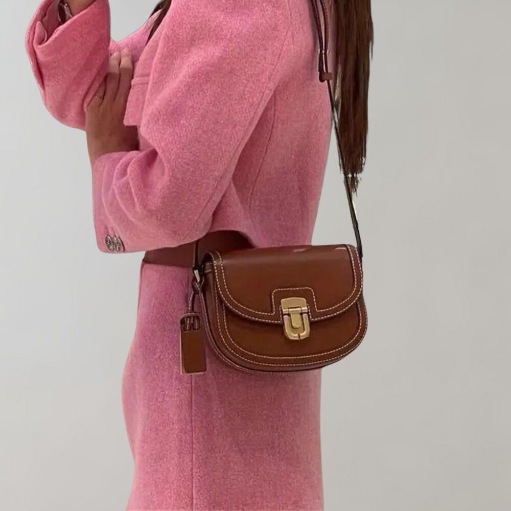 Damenhandtasche aus braunem Leder mit goldener Schnalle, getragen zu einem rosa Blazer. Stilvoll, elegant, modisch, hochwertiges Accessoire.