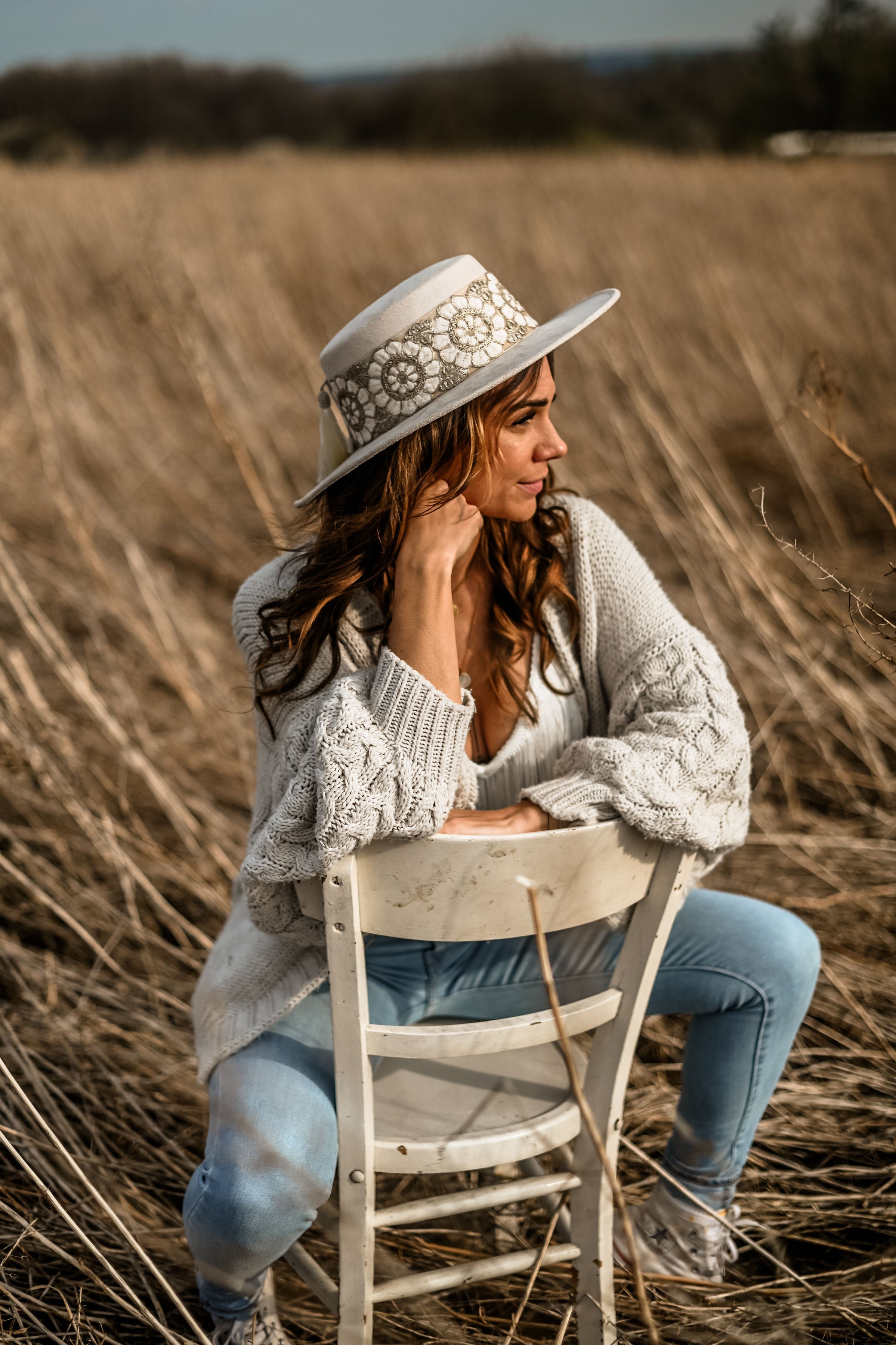 Frau in weißem Hut und Strickjacke sitzt auf Stuhl im Feld. Herbstmode, Outdoor-Fotografie, lässiger Stil, Naturhintergrund.