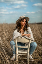 Frau in Jeans und Strickjacke sitzt auf einem Stuhl in einem Feld, trägt Hut, lächelt. Outdoor-Porträt, Natur, Mode, Herbst.