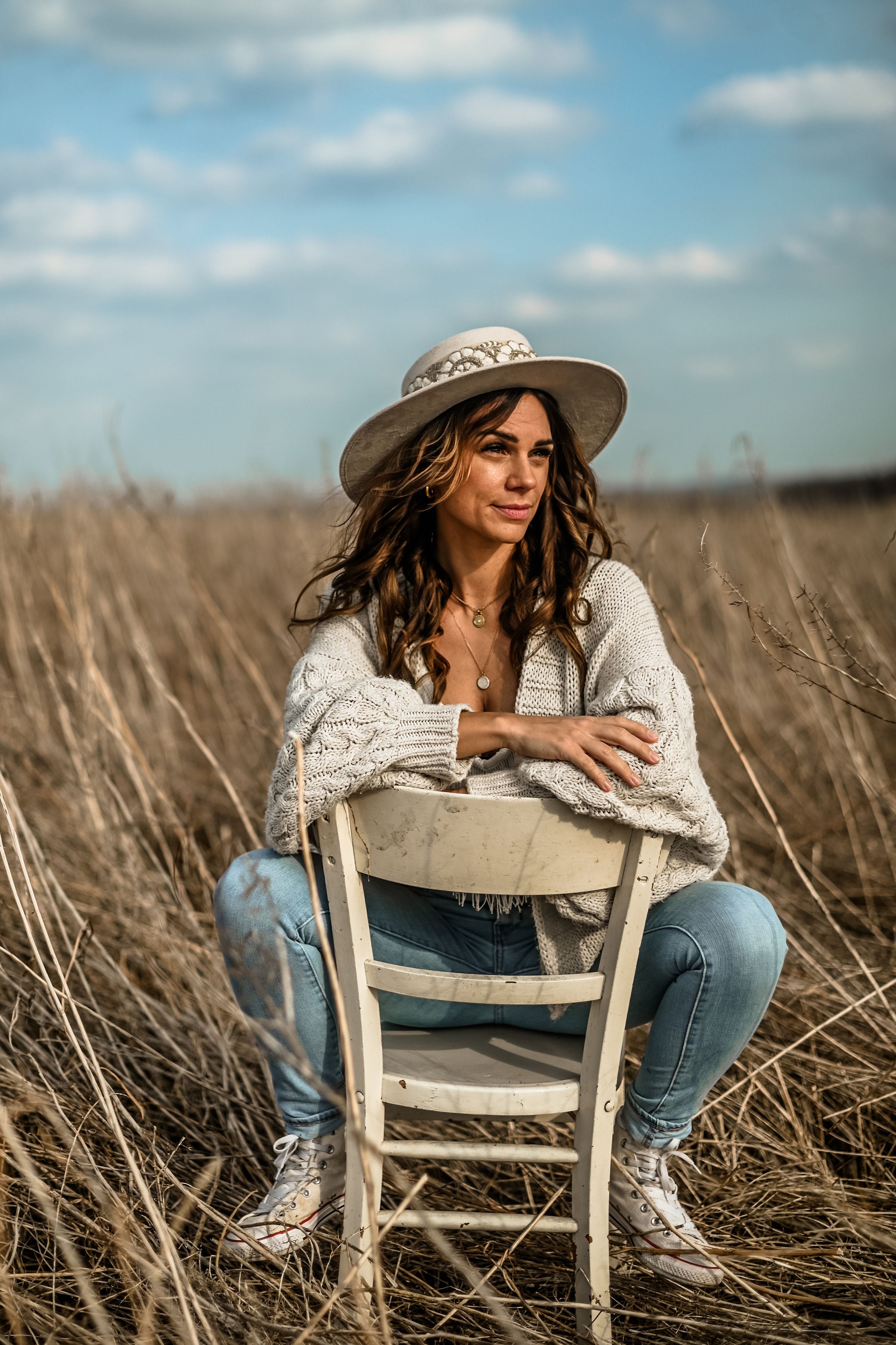 Frau in Jeans und Strickjacke sitzt auf einem Stuhl in einem Feld, trägt Hut, lächelt. Outdoor-Porträt, Natur, Mode, Herbst.