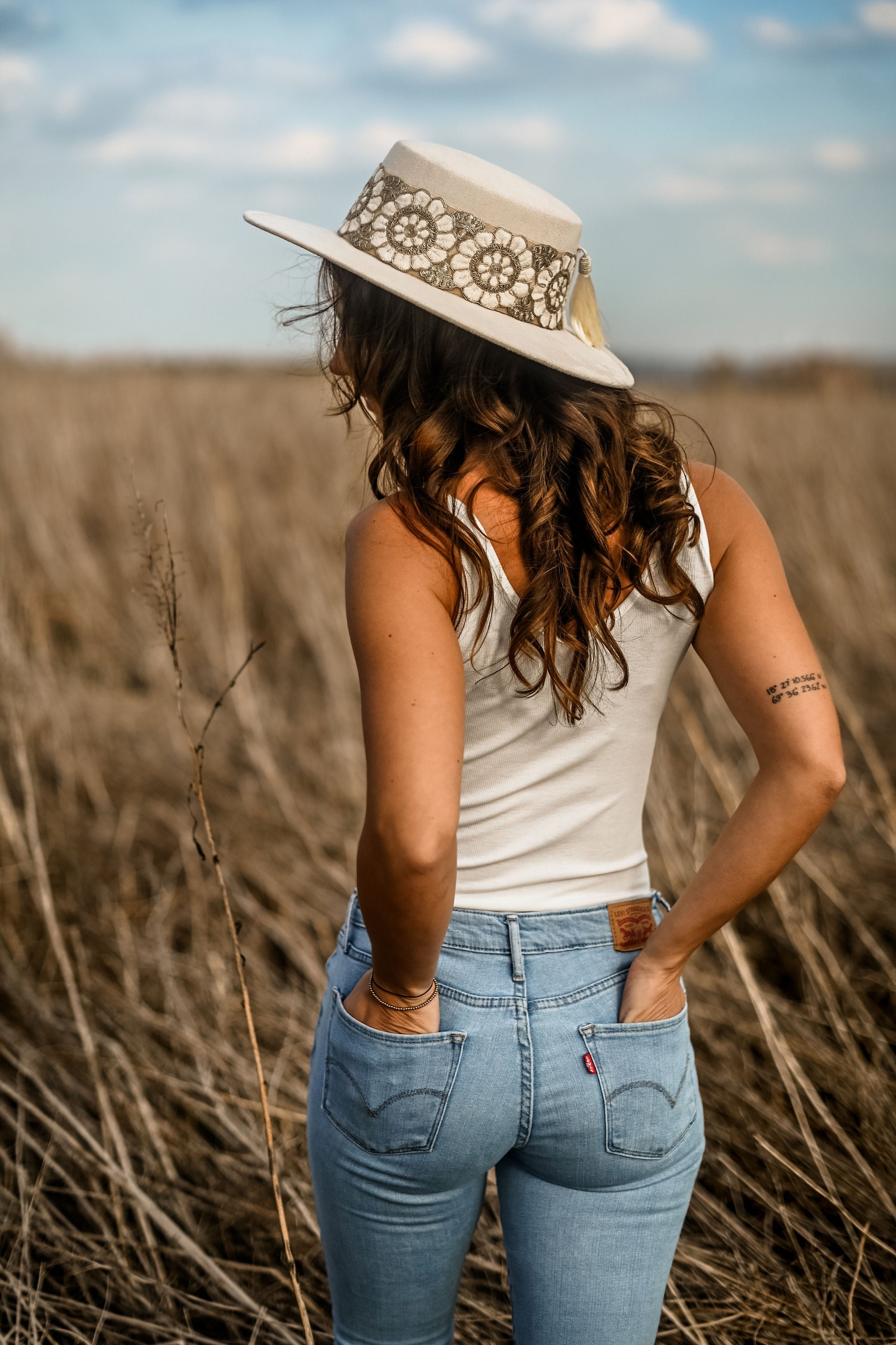 Frau mit weißem Top und Jeans, trägt stilvollen Strohhut mit Spitze, steht in einem Feld. Mode, Sommer, Outdoor, Freizeitkleidung, Strohhut.
