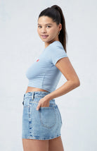 Frau in hellblauem Crop-Top und Jeansrock, lächelt seitlich. Modetrend, Sommeroutfit, lässiger Stil, Damenmode, Freizeitkleidung.