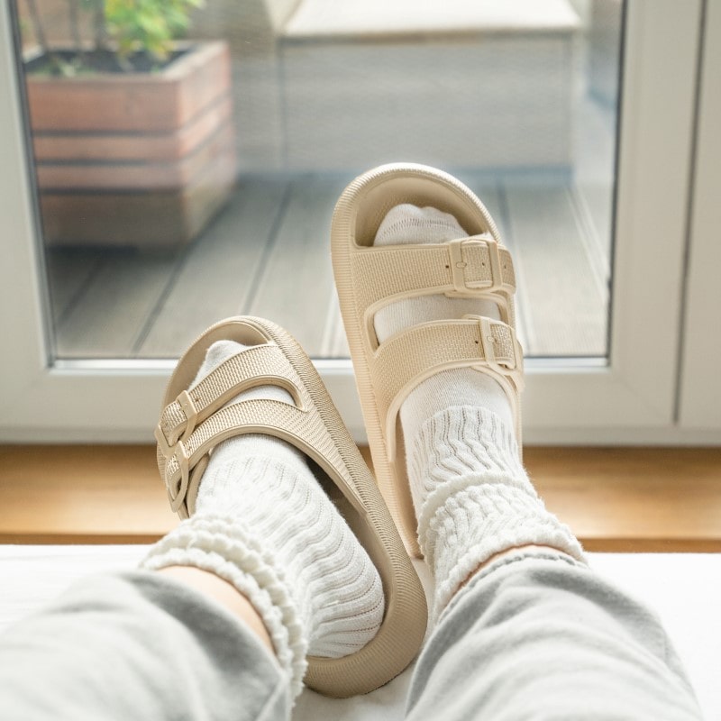 Beige Sandalen mit Doppelriemen, getragen mit weißen Socken, vor einem Fenster. Bequeme Hausschuhe, ideal für den Alltag und Freizeit.