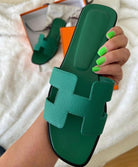 Grüne Ledersandale mit markantem H-Design, gehalten von einer Hand mit grünen Nägeln. Luxuriöse Sommerschuhe, stilvoll und modern.