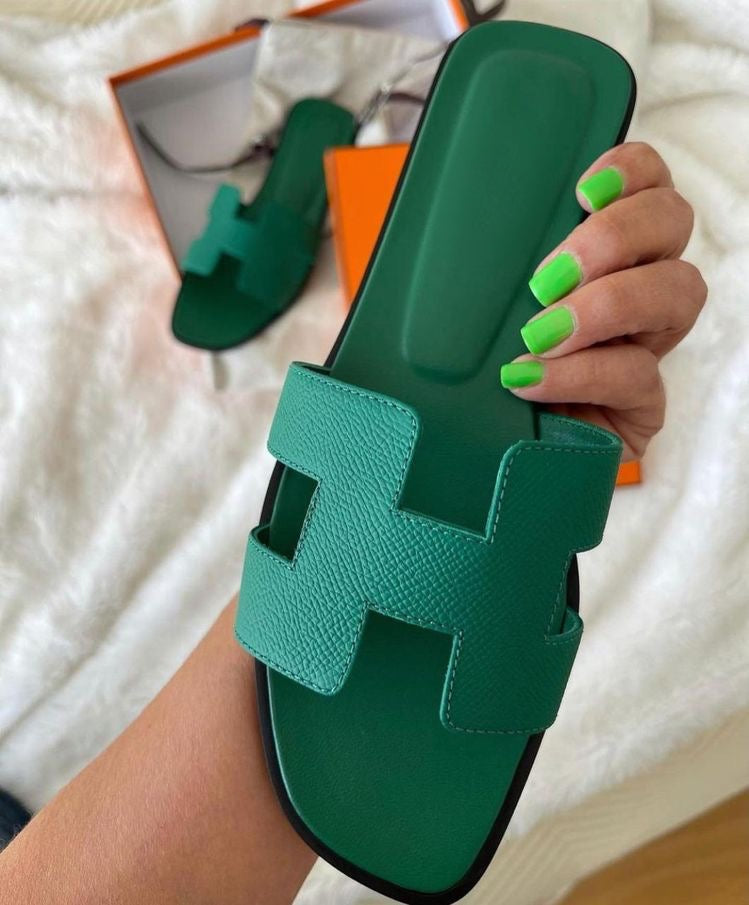 Grüne Ledersandale mit markantem H-Design, gehalten von einer Hand mit grünen Nägeln. Luxuriöse Sommerschuhe, stilvoll und modern.