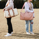 Zwei Personen mit faltbaren Reisetaschen, eine in Weiß und Braun, die andere in Rosa, auf einem Feld.