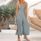 Frau in grauem Jumpsuit, Sommermode, Strand, Strohtasche, Pflanzen im Hintergrund.