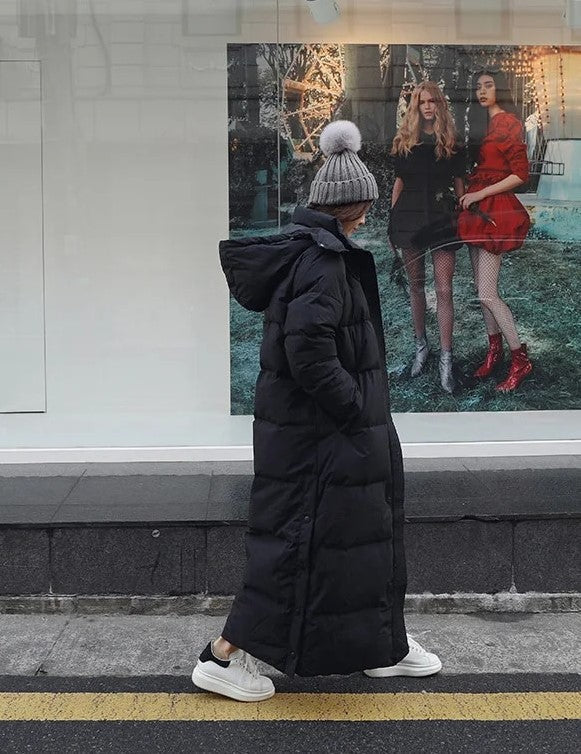 Langer schwarzer Daunenmantel mit Kapuze, getragen von Person auf Straße. Wintermode, warm, stilvoll, urbaner Look, Streetwear-Trend.