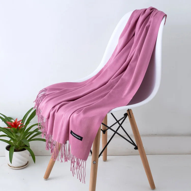 Rosa Kaschmirschal auf weißem Stuhl, weiche Fransen, luxuriöses Accessoire, ideal für Herbstmode, stilvoll und elegant, neben grüner Pflanze.