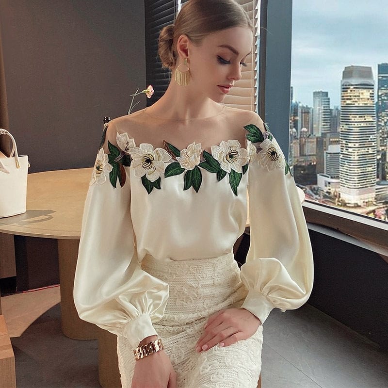 Elegante Frau in weißer Bluse mit Blumenstickerei, sitzt vor Fenster mit Stadtblick. Mode, Damenbekleidung, stilvolle Bluse, urbaner Chic.
