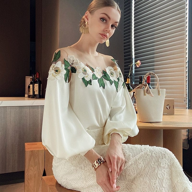 Elegantes Damenoutfit mit weißer Bluse, floralen Stickereien und langen Ärmeln. Perfekt für stilvolle Anlässe und modische Events.
