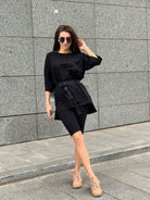 Frau in schwarzem Oversize-T-Shirt und Radlerhose, kombiniert mit beigen Sneakers, posiert vor grauer Wand. Streetwear, Mode, urbaner Stil.