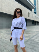 Frau in weißem Oversize-Shirt und Radlerhose, trägt Sonnenbrille, steht vor modernem Gebäude. Streetstyle, urbaner Look, Mode 2023.