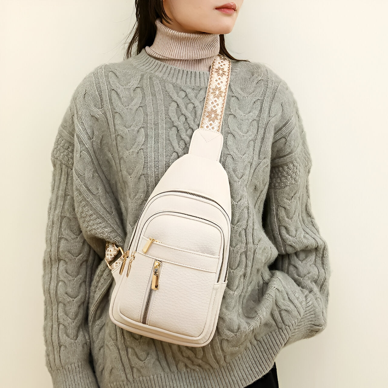 Frau mit grauem Strickpullover und beiger Umhängetasche mit goldenen Reißverschlüssen.