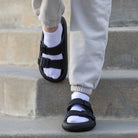 Schwarze Sandalen mit verstellbaren Riemen, getragen mit weißen Socken und grauer Jogginghose auf Treppe. Bequeme, modische Fußbekleidung.