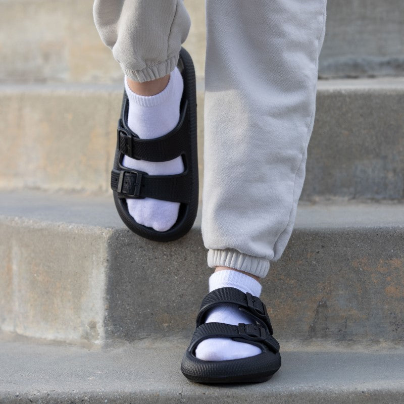 Schwarze Sandalen mit verstellbaren Riemen, getragen mit weißen Socken und grauer Jogginghose auf Treppe. Bequeme, modische Fußbekleidung.