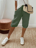 Frau in grüner Leinenhose, weißem T-Shirt, weißen Sneakers, beiger Umhängetasche im Wohnzimmer.