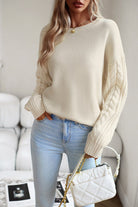 Damenpullover in Creme mit Zopfmuster-Ärmeln, kombiniert mit hellblauer Jeans und weißer Umhängetasche. Elegante Herbstmode für Frauen.