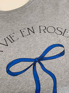 Graues T-Shirt mit blauem Schleifenmotiv und "VIE EN ROSE" Schriftzug. Modisches Design, hochwertiger Druck, trendige Damenbekleidung.