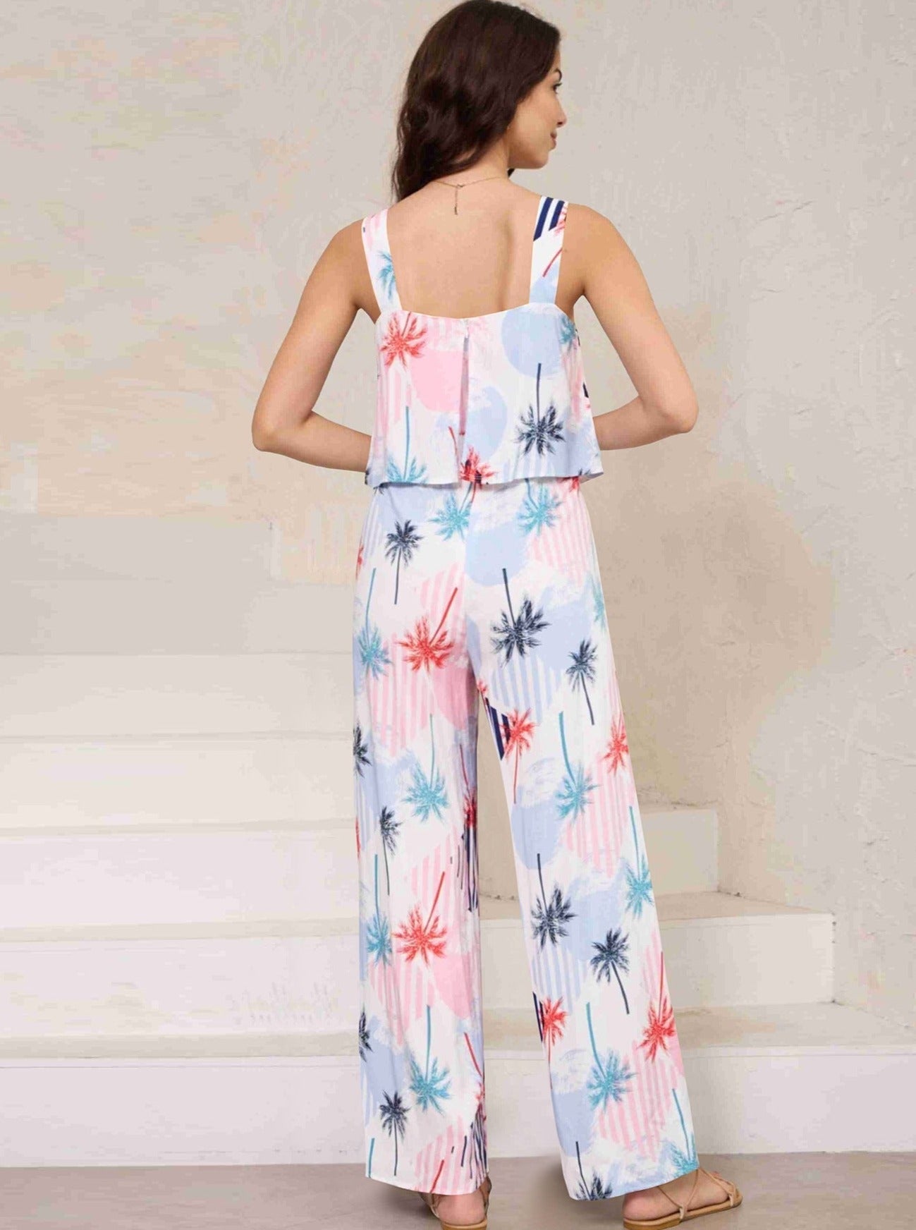 Frau in sommerlichem Jumpsuit mit Palmenmuster, ärmellos, pastellfarben, ideal für Strandmode und Freizeitkleidung. Perfekt für warme Tage.