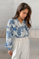 Elegante Frau in weißer Bluse mit blauem Toile-de-Jouy-Muster, kombiniert mit stilvollen, weißen Hosen. Perfekt für modische Büro-Outfits.