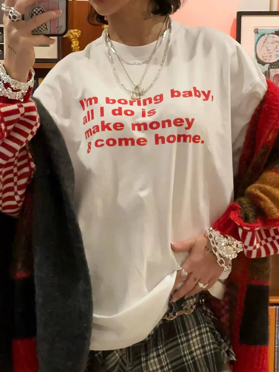 Person trägt ein weißes T-Shirt mit rotem Text "I'm boring baby, all I do is make money & come home", kombiniert mit karierten Hosen und Schmuck.