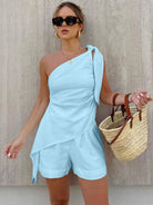 Frau in hellblauem, schulterfreiem Jumpsuit mit Strohtasche, Sonnenbrille und goldenen Accessoires. Sommermode, eleganter Look, Damenbekleidung.