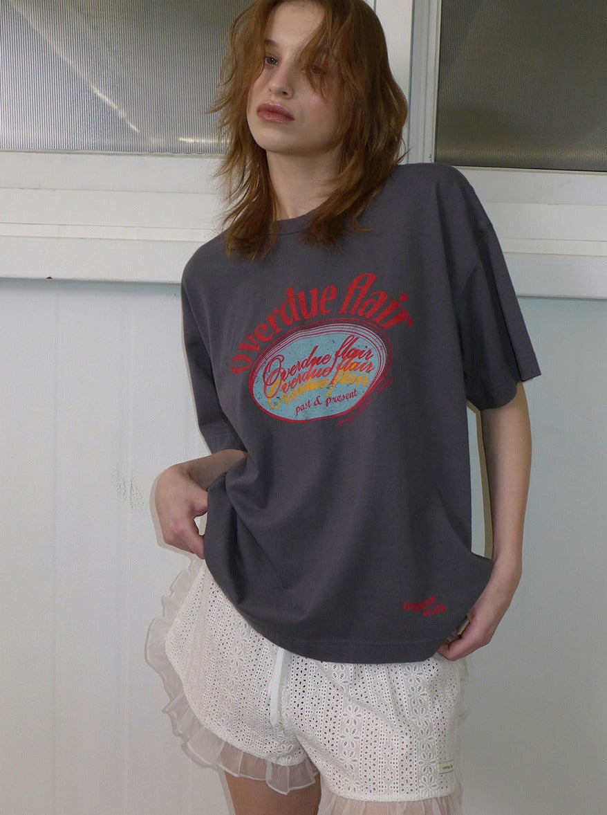 Frau trägt graues T-Shirt mit rotem "Overdue Flair"-Aufdruck und weiße Spitzen-Shorts. Modetrend, lässiger Stil, Streetwear, Damenmode.