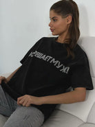 Frau in schwarzem T-Shirt mit auffälligem Schriftzug, sitzt entspannt auf einem weißen Sofa. Modetrend, lässiger Look, Freizeitkleidung.