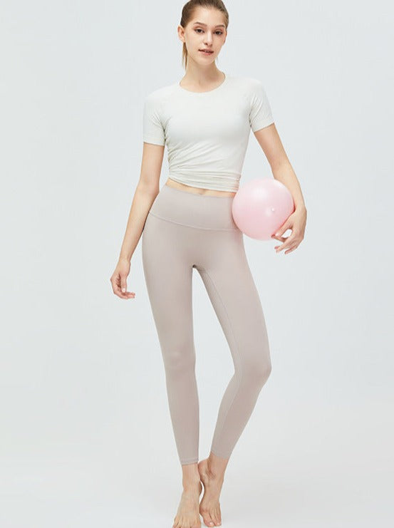 Frau in beiger Leggings und weißem T-Shirt hält rosa Gymnastikball. Sportbekleidung, Fitnessmode, Yoga-Outfit, athletische Kleidung.