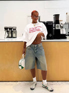 Frau in lässigem Outfit mit weißem T-Shirt und Jeansshorts, steht vor einer modernen Kaffeebar. Trendige Mode, urbaner Stil, Streetwear.