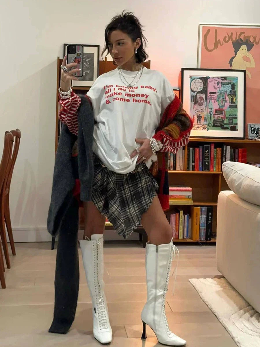 Frau in modischem Outfit mit weißem T-Shirt, kariertem Rock und weißen Stiefeln posiert in stilvollem Wohnzimmer. Trendige Damenmode, urbaner Look.