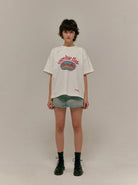 Weißes Oversize-T-Shirt mit "Overdue Flair"-Aufdruck, kombiniert mit Jeansshorts und schwarzen Stiefeln. Modetrend, lässiger Streetstyle.