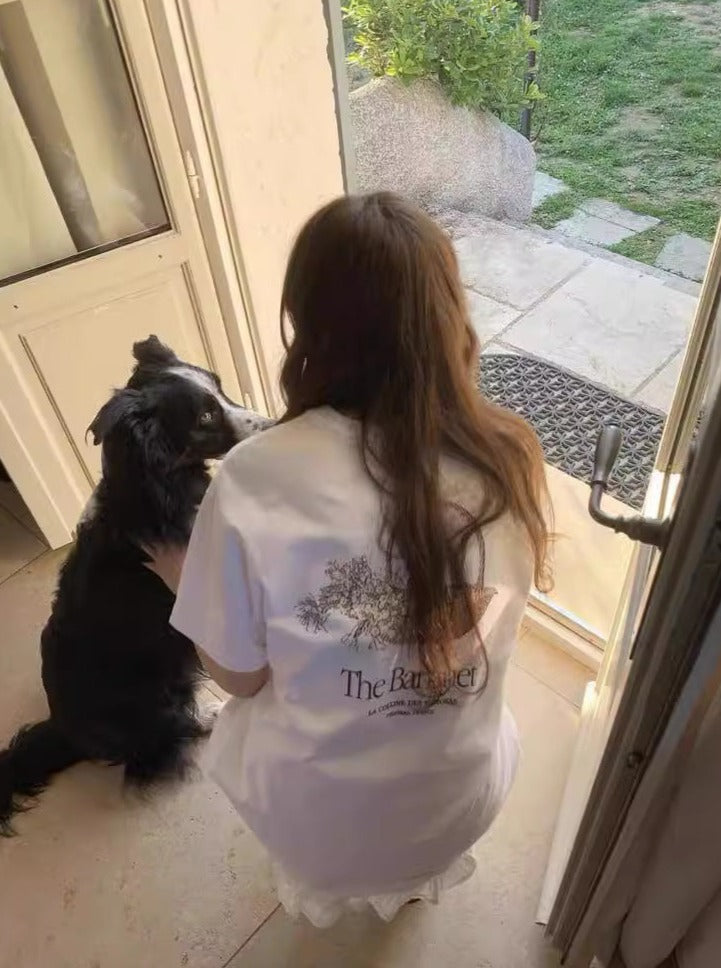 Frau in weißem T-Shirt mit Hund an der Tür, Gartenblick. Keywords: Frau, Hund, T-Shirt, Tür, Garten, Haustier, Natur, Entspannung.
