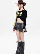 Frau in modischem Outfit: schwarzes Langarmshirt mit Schmetterlingsmotiv, grauer Minirock, Cowboystiefel, brauner Hut. Trendige Streetwear.